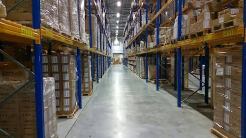 Spacious modern warehouse interior in Dubai with organized pallets of boxes on blue and orange metal shelving racks along a clean, brightly lit aisle.