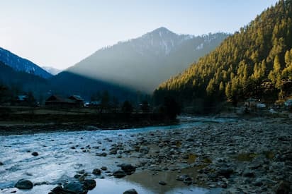 cenic river valley in Srinagar, Kashmir, with rocky stream, forested mountains, snow-capped peaks, and traditional houses at dawn.