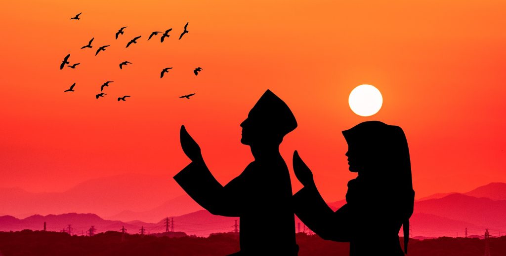 Silhouette of a Muslim man and woman in traditional attire praying with hands raised against a vibrant orange sunset, with birds flying overhead and purple mountains in the background.