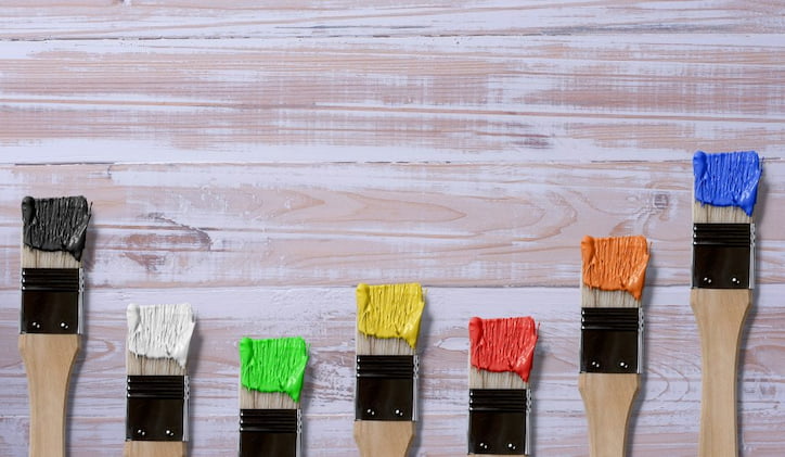 Seven paintbrushes dipped in multicolored paints on a light wooden background