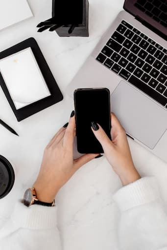 Top-down view of a modern workspace with a hand holding a smartphone, alongside a laptop, blank notebook, pen holder, pencil, coffee cup, and watch on a white marble desk