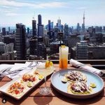 Rooftop dining scene featuring tacos and a cocktail with a panoramic view of the Toronto skyline, including the CN Tower, under a partly cloudy sky.