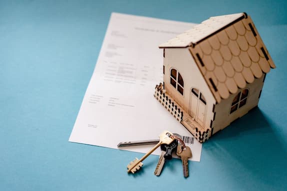 A wooden model of a house placed on a blank contract document, accompanied by a set of keys with a barcode tag, all set against a light blue background.